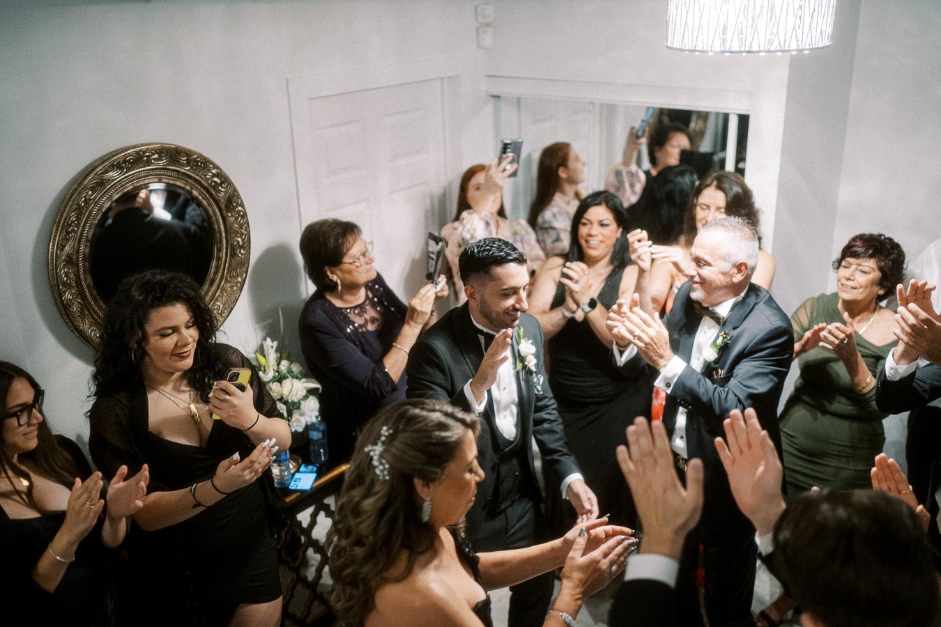 Elegant wedding celebration with guests clapping and taking photos, featuring a smiling groom in a black tuxedo surrounded