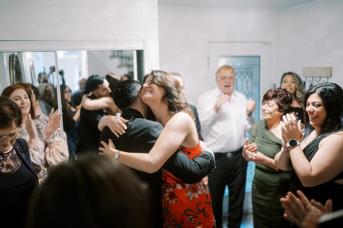 A joyful group celebrates as a woman in a red floral dress hugs a man in a suit, surrounded by clapping and smiling people