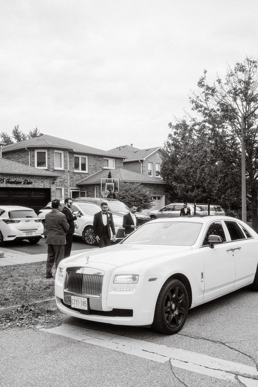 Monochrome image of a luxury neighborhood scene featuring a white Rolls-Royce parked on the street. Several people