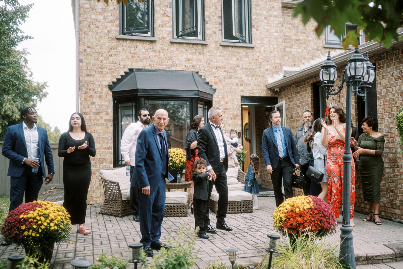 A group of well-dressed people gathering in a brick patio area for an outdoor event, with colorful flowers and a brick