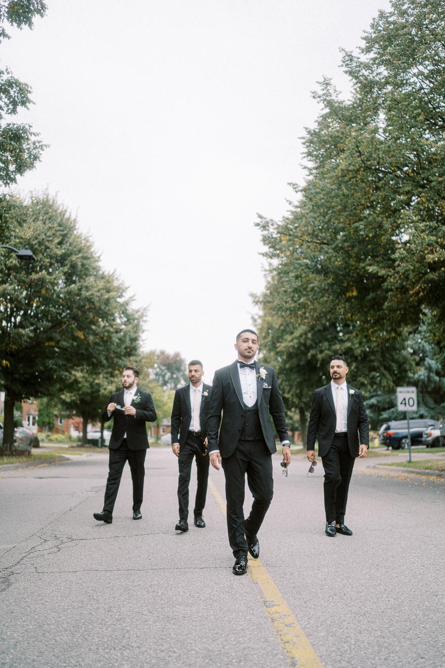Four men in formal black suits and bow ties walk confidently down a tree-lined street, showcasing elegant wedding attire.
