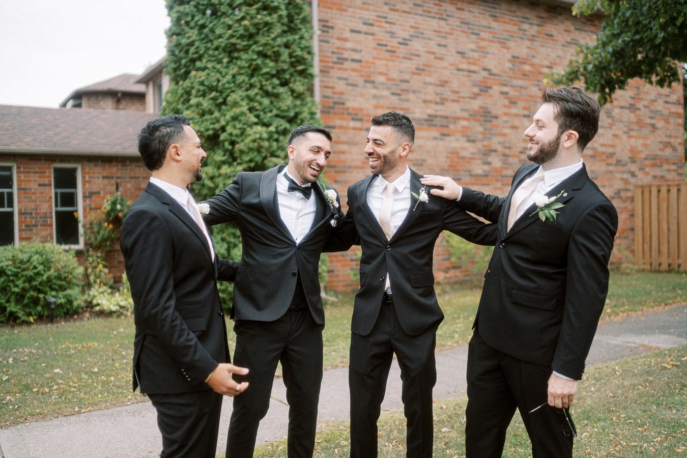 A group of four groomsmen in black suits and white shirts smiling and embracing each other outdoors, in front of a brick
