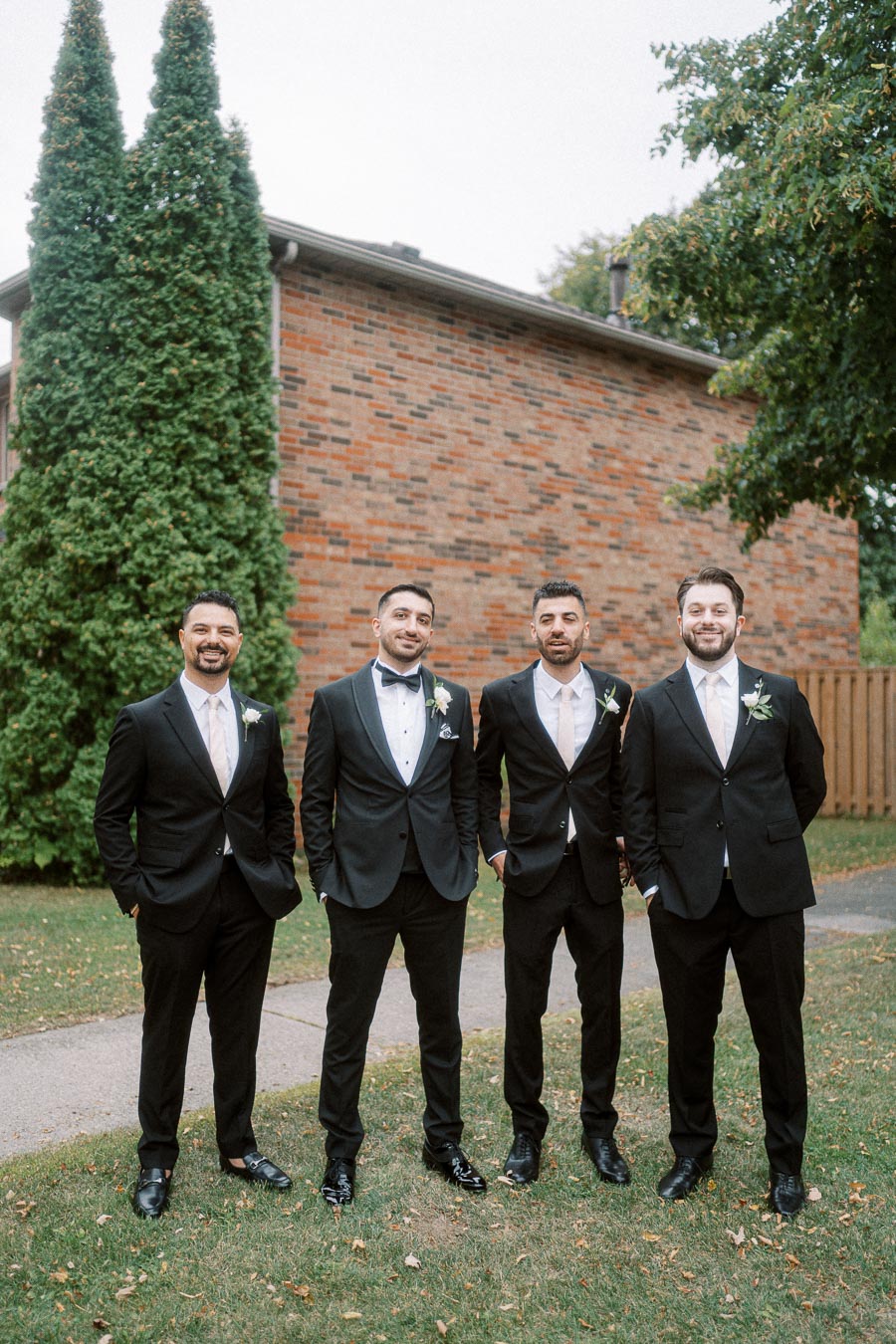 Four groomsmen in black suits pose outdoors near a brick building and greenery, dressed for a wedding, exuding elegance and