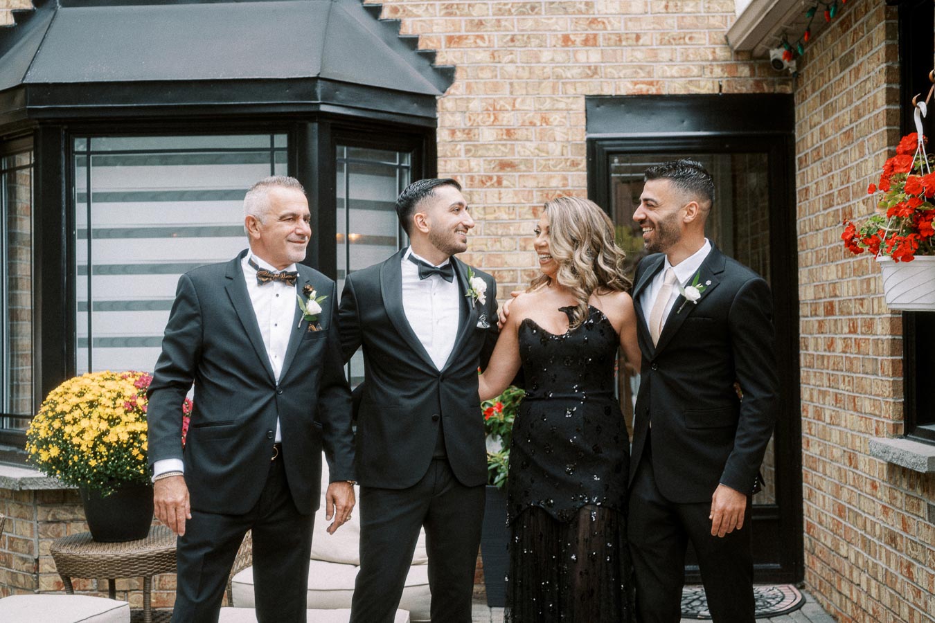 A group of four people dressed in formal attire, including three men in tuxedos and a woman in a black evening gown, smiling