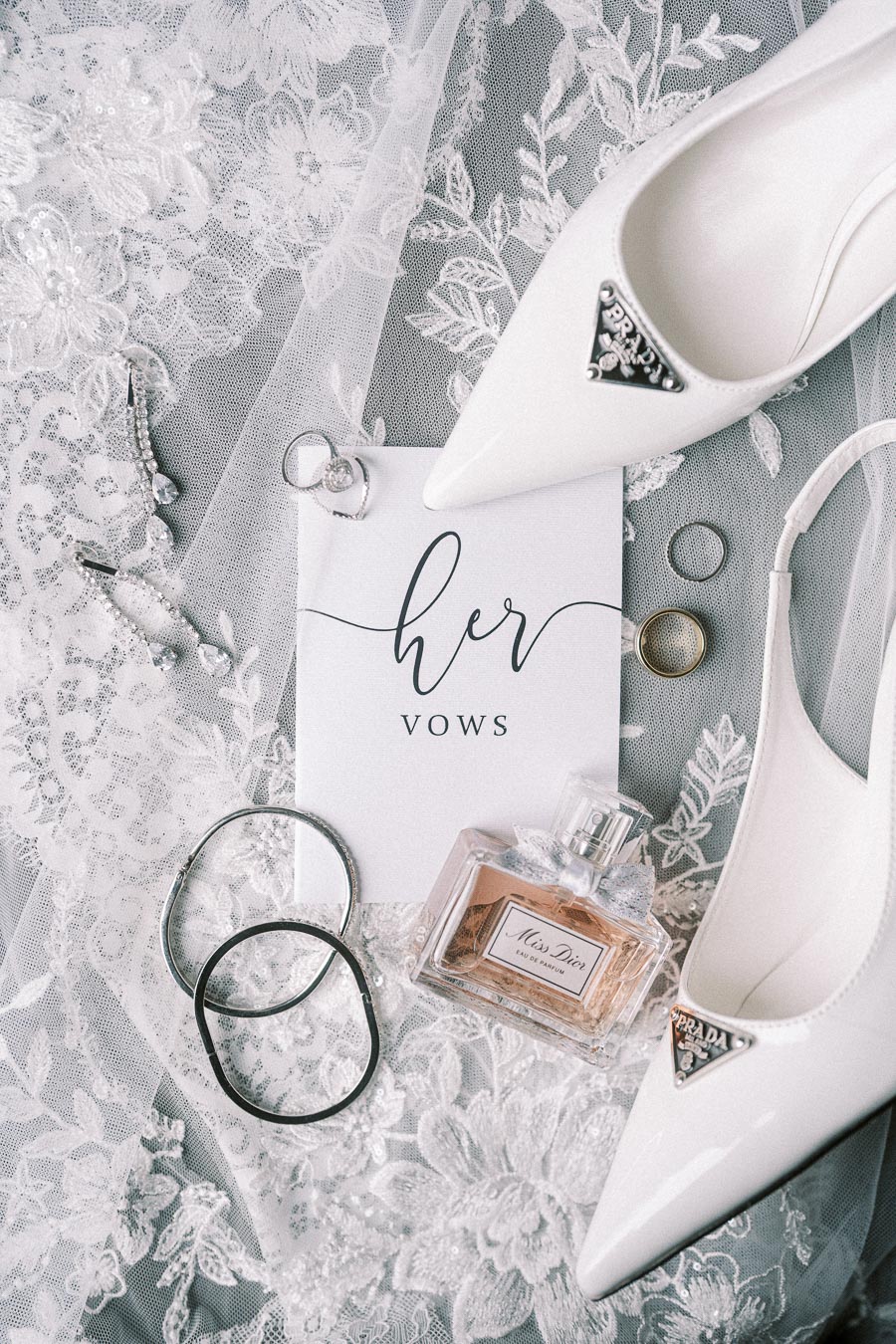 Bridal accessories including white high heels, perfume, wedding rings, jewelry, and a vow card displayed on delicate lace