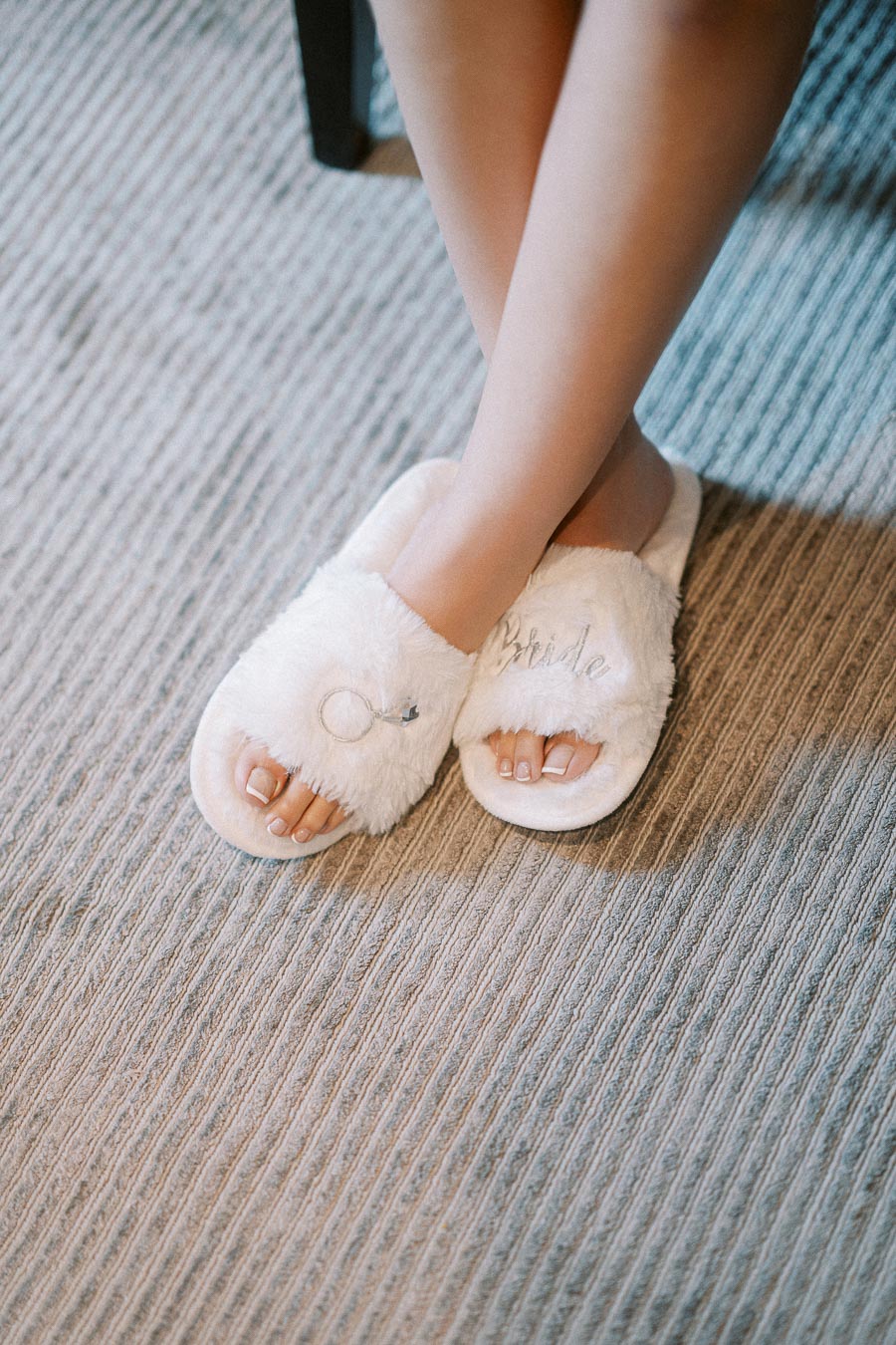 Close-up of a woman's legs crossed, wearing cozy white bridal slippers with 'Bride' and engagement ring embroidery, resting