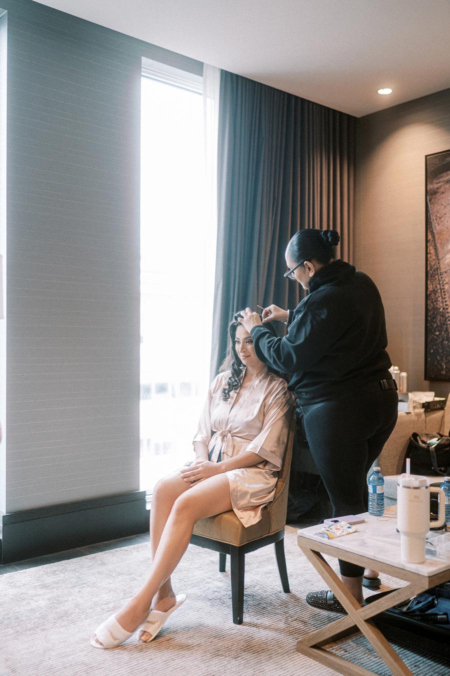 Woman getting ready for an event as a hairstylist works on her hair in a hotel room with natural light coming through the
