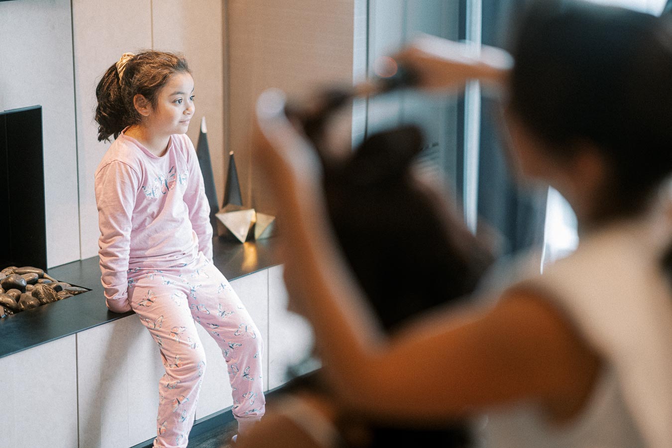 Young girl in pink pajamas sitting on a modern fireplace hearth, with blurred person adjusting camera equipment in the