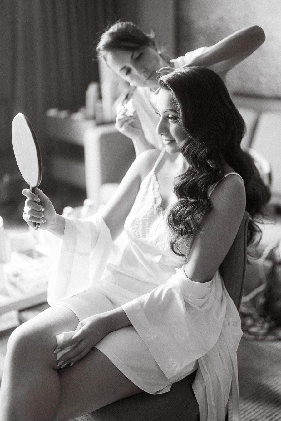 Bridal preparation scene with woman in silk robe having her hair styled while smiling at her reflection in a hand mirror.