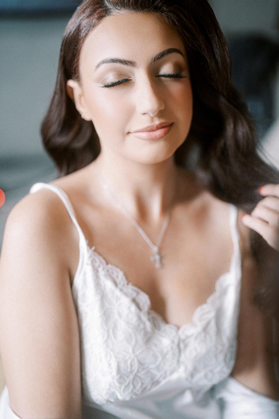 A woman with long, dark hair and closed eyes wearing a white lace camisole and a silver necklace with a cross pendant,