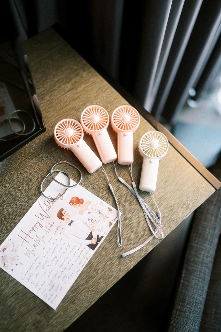 Four portable mini fans in shades of pink and white on a wooden table next to a wedding card with a bride and groom