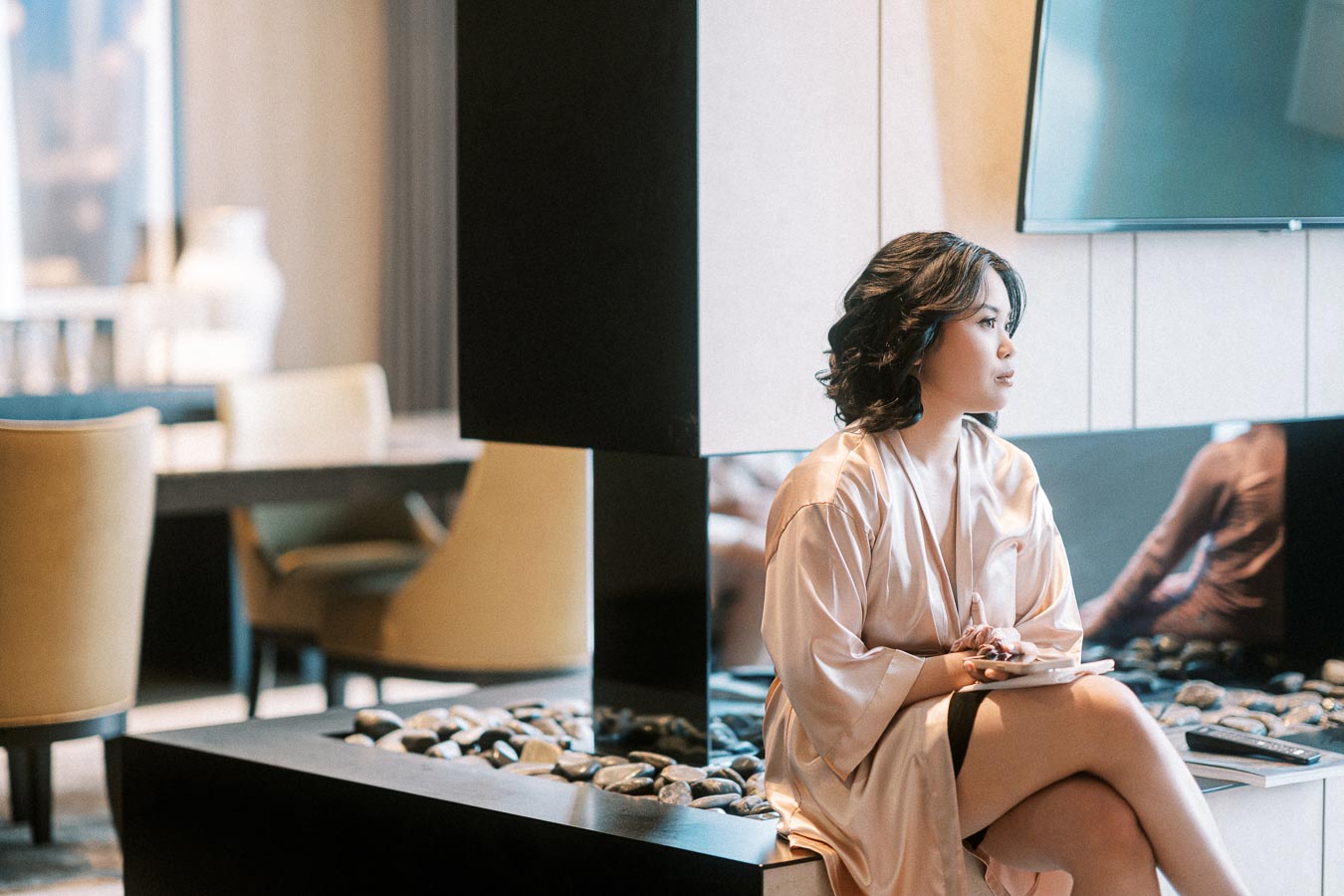 Elegant woman in a silk robe sitting in a modern, stylish living area with neutral tones and a contemporary fireplace,