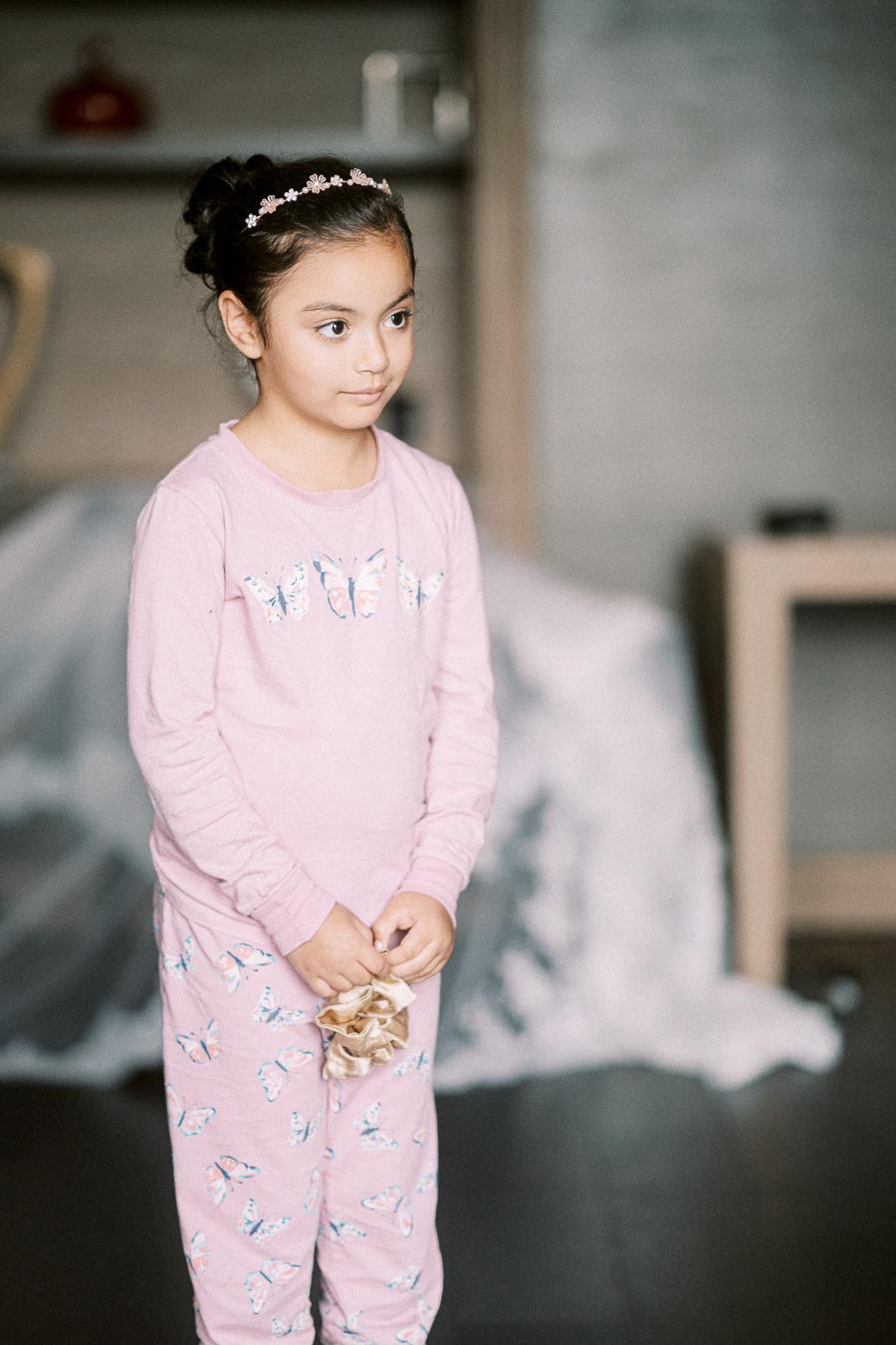 A young girl wearing a pink butterfly-patterned pajama set and a floral headband stands indoors, holding a scrunchie, with a