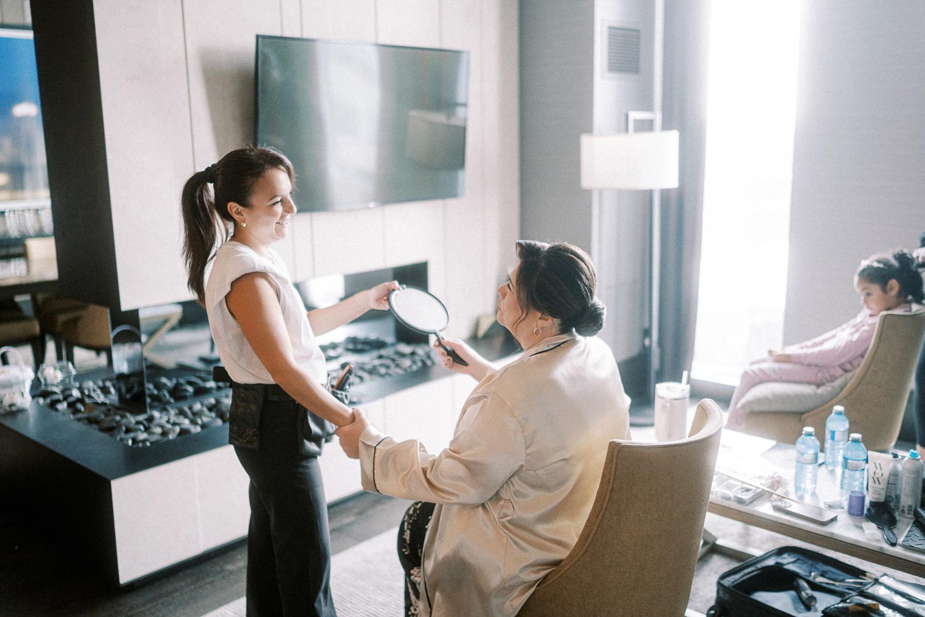 A woman in a comfortable room getting her makeup done by a makeup artist holding a hand mirror, with a young girl sitting