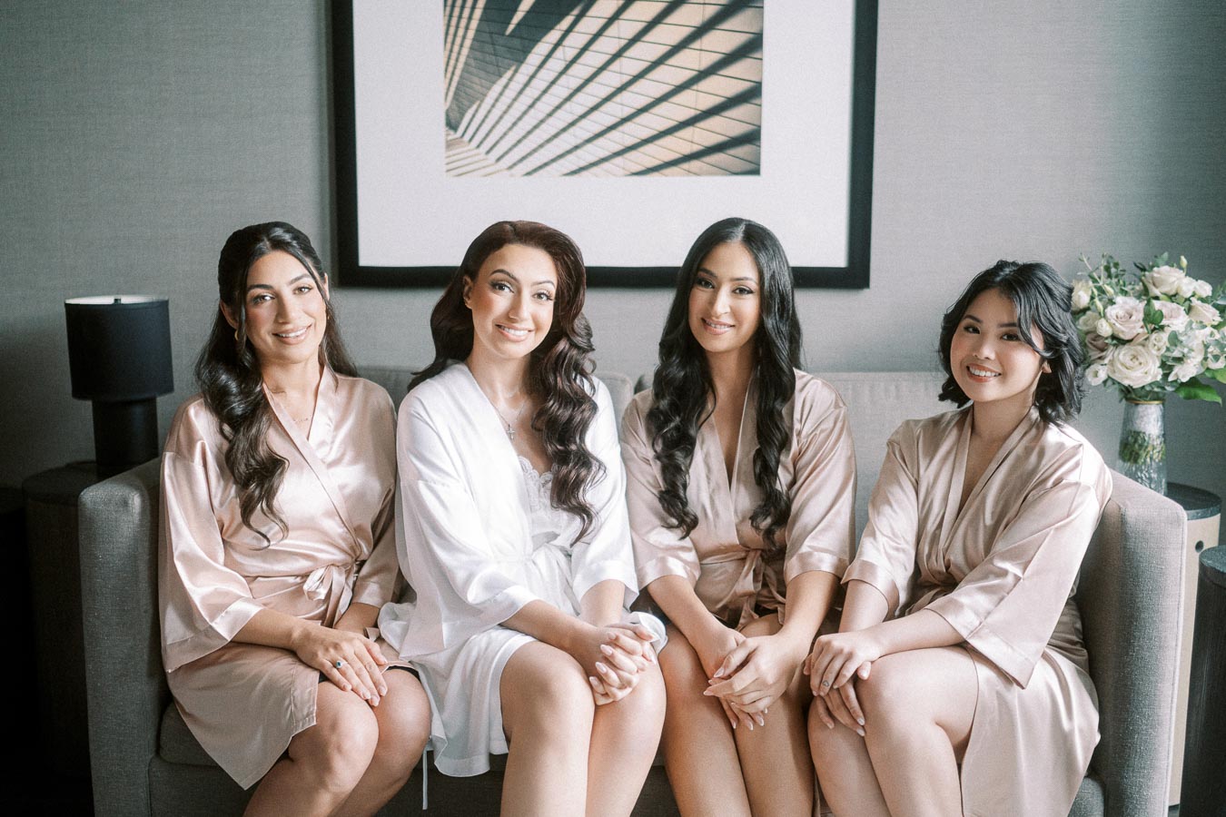 A group of four women in matching silk robes sitting on a sofa, smiling, indoors, with a modern art piece and a bouquet of