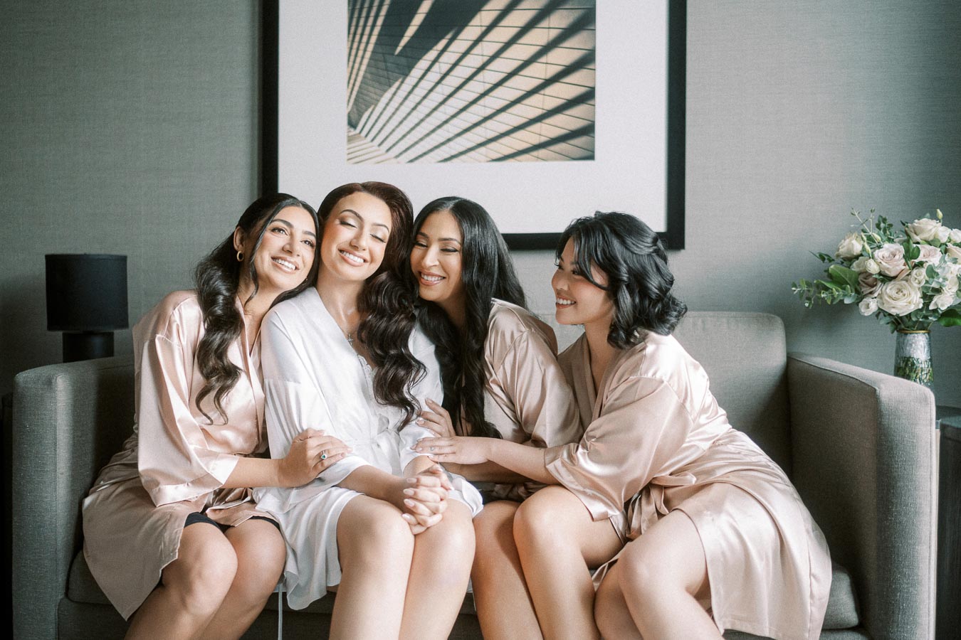 A group of four women in matching silk robes sitting on a sofa, smiling and embracing, with a modern artwork and bouquet of