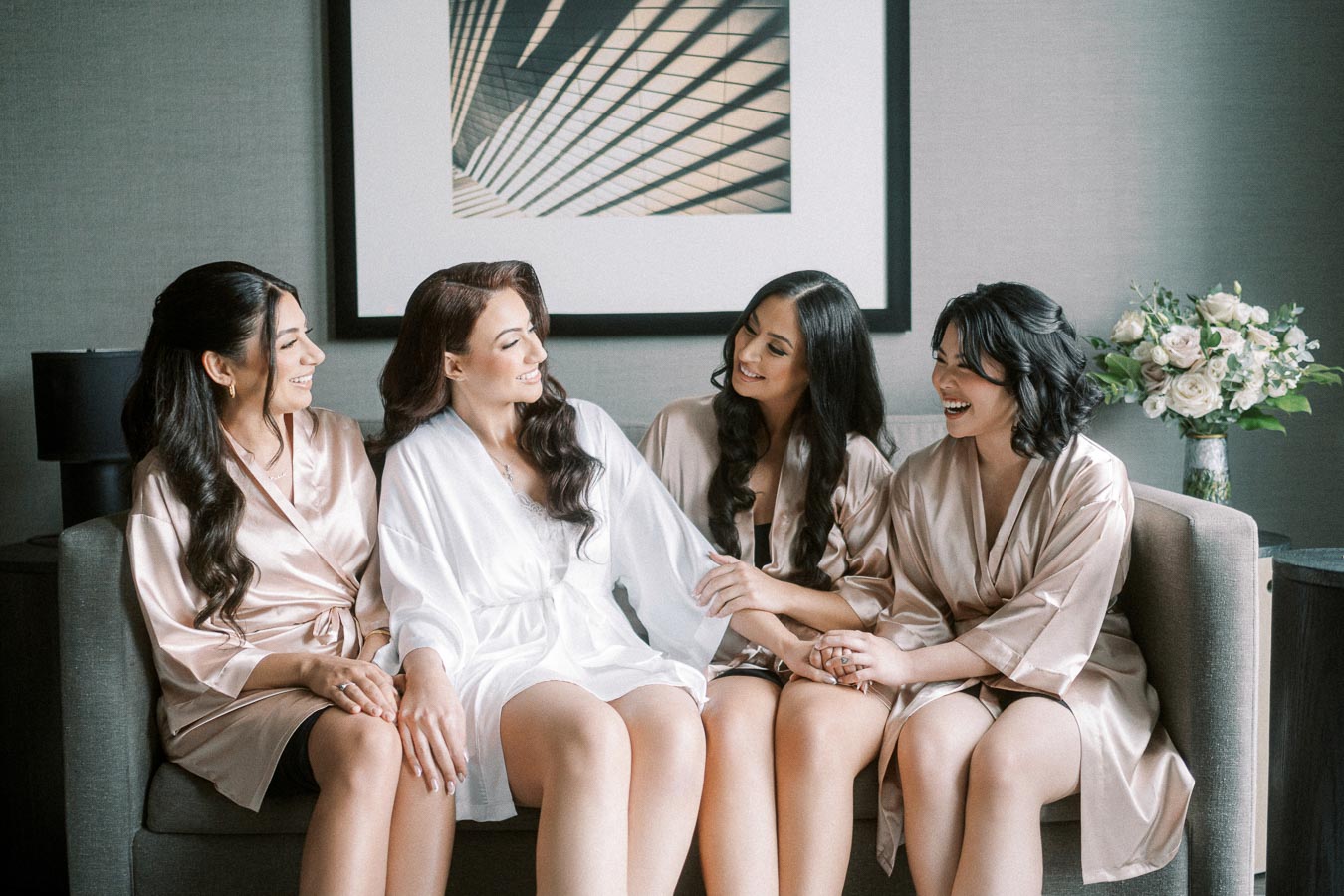 Four women in silk robes sit together on a couch, happily chatting and laughing. The setting is a cozy room with a modern