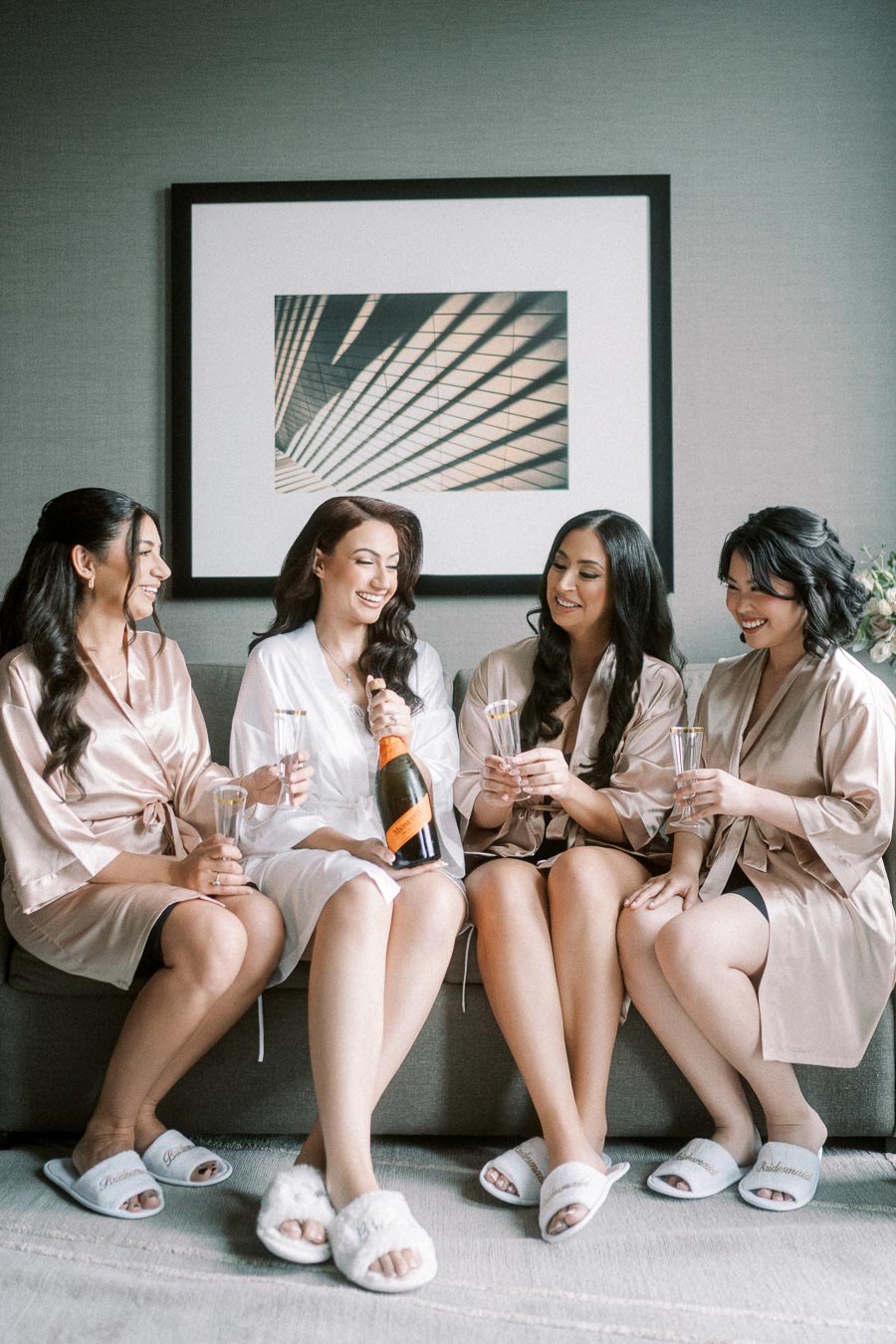 Four women in silk robes sit on a couch, smiling and holding glasses, while celebrating with champagne, in a modern, chic