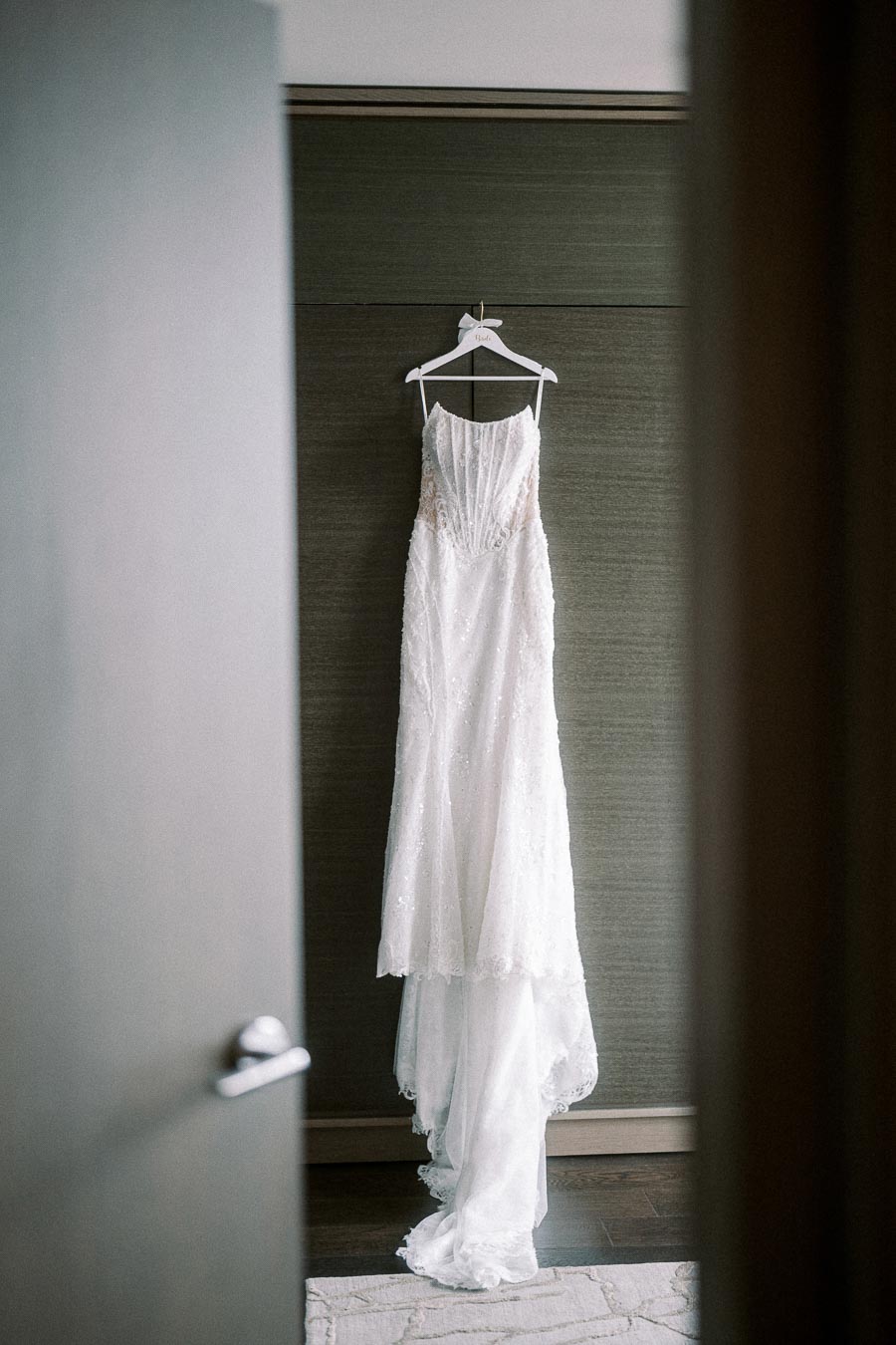 Elegant white wedding dress hanging on a wooden hanger in a softly lit room, showcasing intricate lace details and a flowing