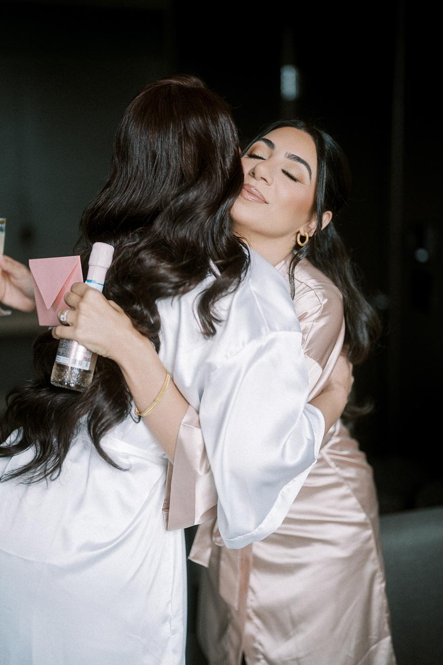 Two women in satin robes embrace tightly, sharing a joyful moment. One holds a small bottle and card, suggesting a