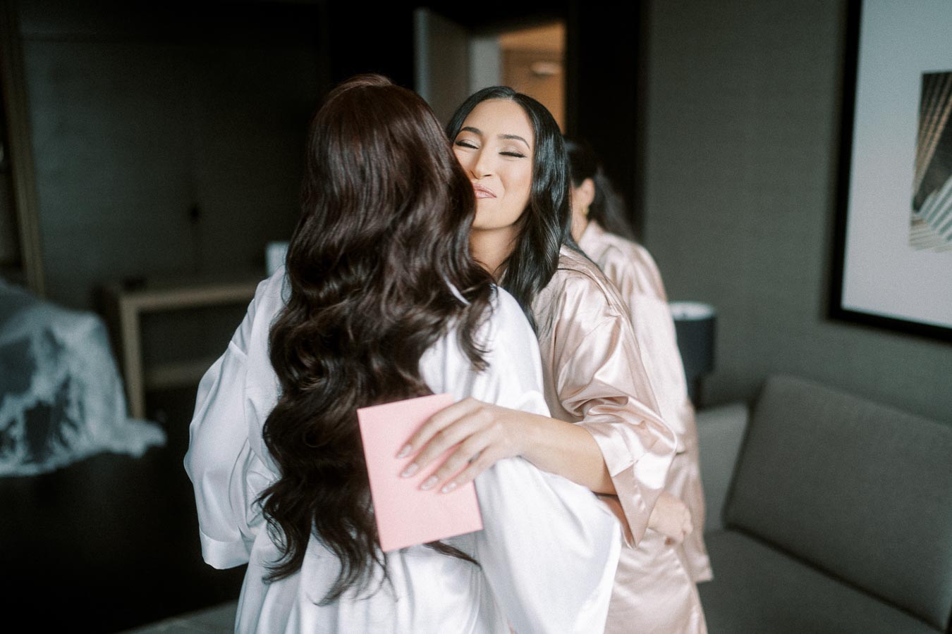 Two women in silk robes hugging in a cozy room, holding a card, creating a warm and affectionate atmosphere.