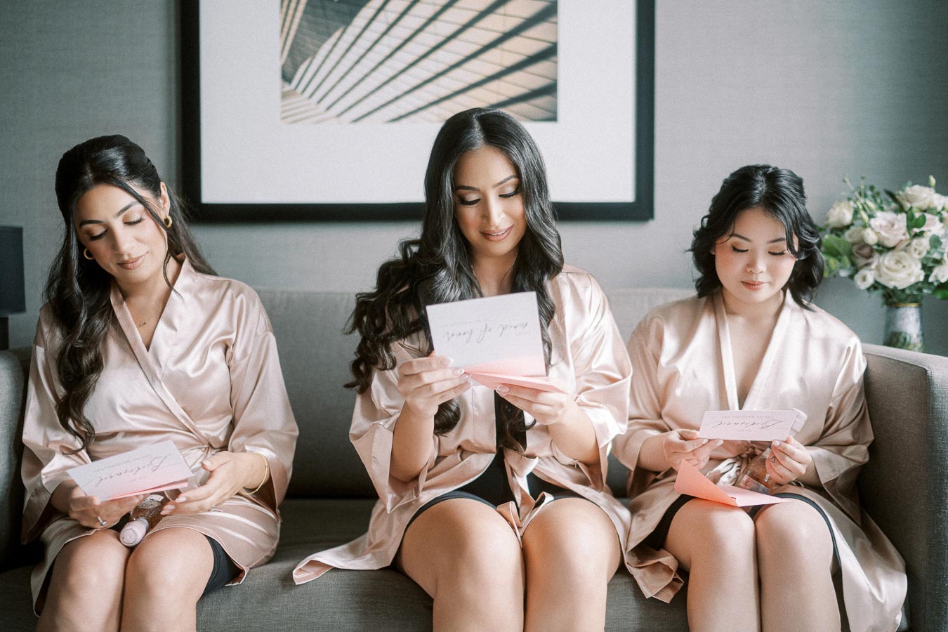 Three women in matching satin robes sit on a sofa, reading cards with smiles. A bouquet of flowers is visible in the