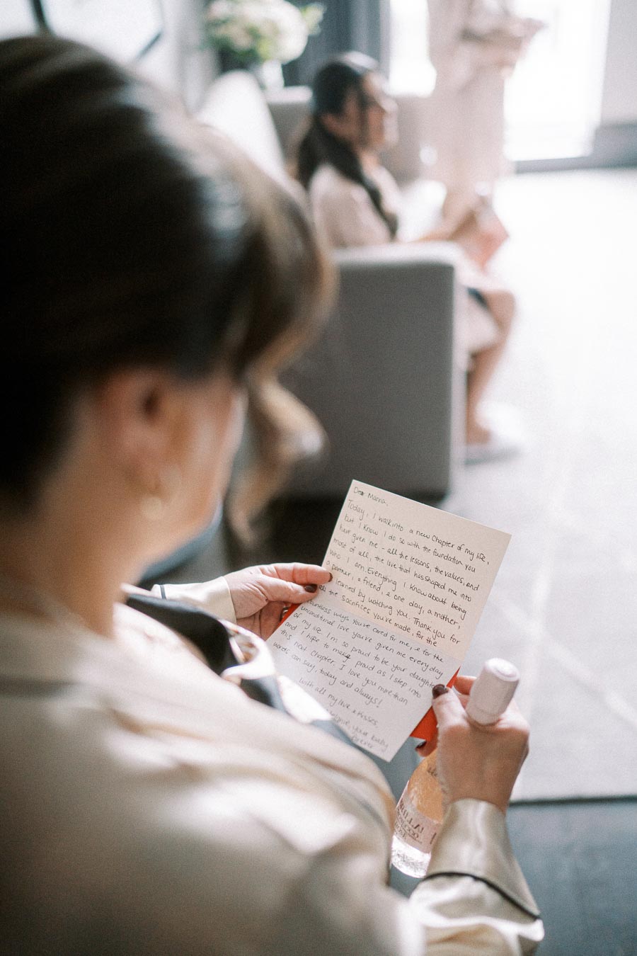 A person wearing a satin robe holds a handwritten letter, likely on a wedding day. In the background, another person in a