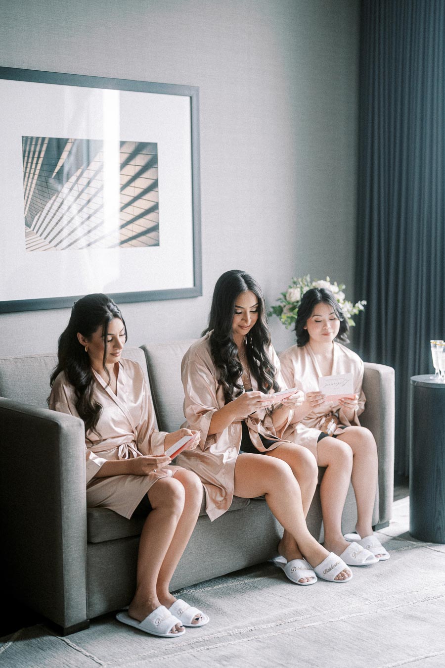 Three women wearing matching silk robes and slippers sit on a sofa, reading cards in a stylish room with modern decor.