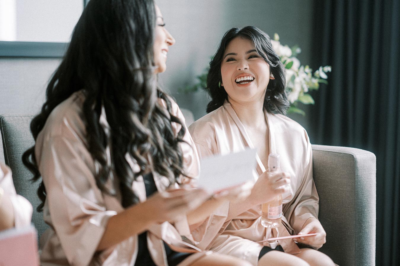 Two women in silk robes laughing and enjoying a relaxed moment, holding cards or letters, with a soft focus background