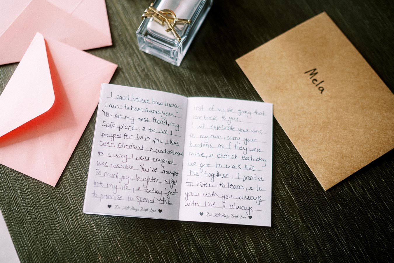 Handwritten love letter with a pink envelope and perfume bottle on a wooden table, next to a brown envelope labeled Mela.
