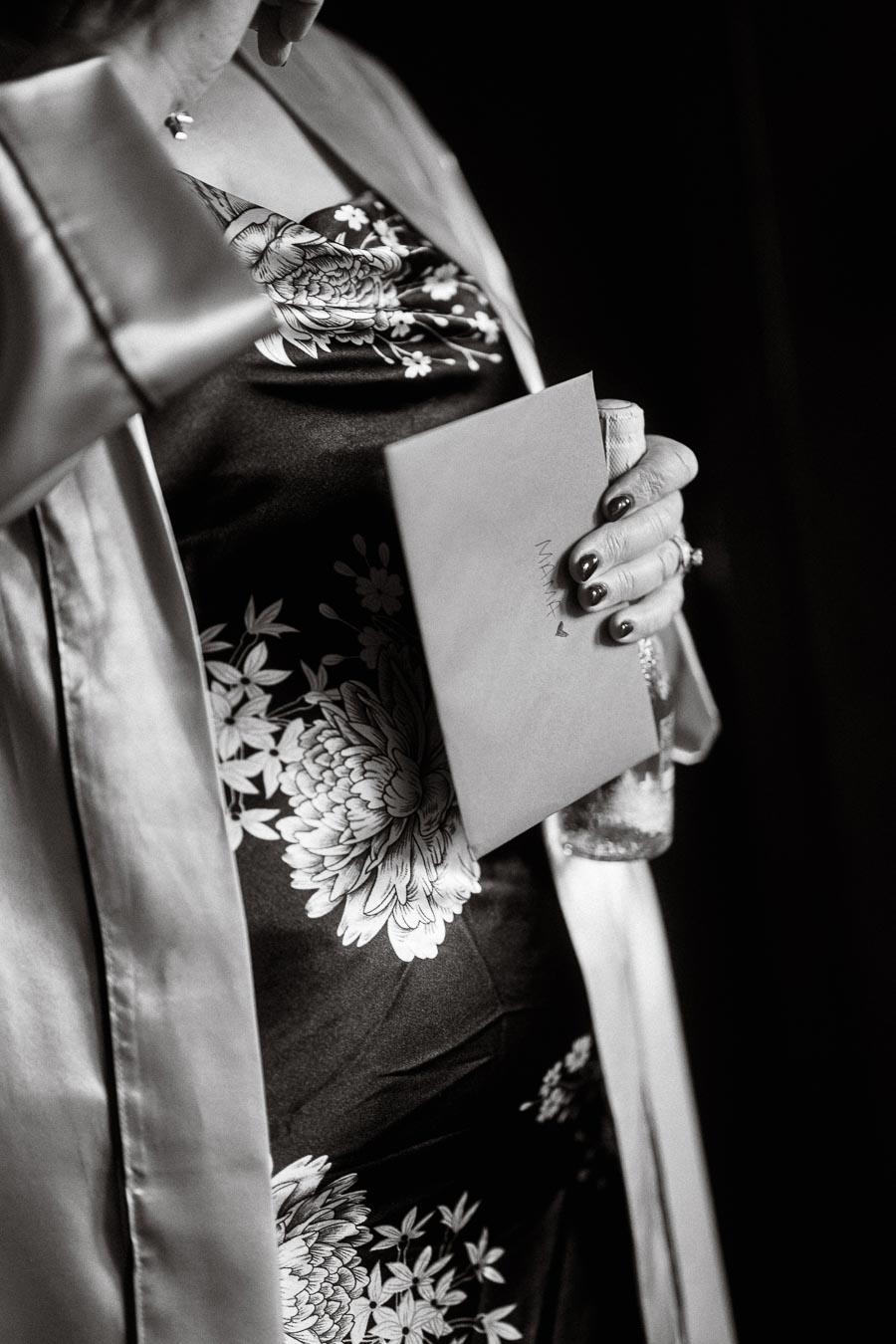Black and white close-up of a person holding a card with Mama written on it, wearing a floral patterned dress and satin