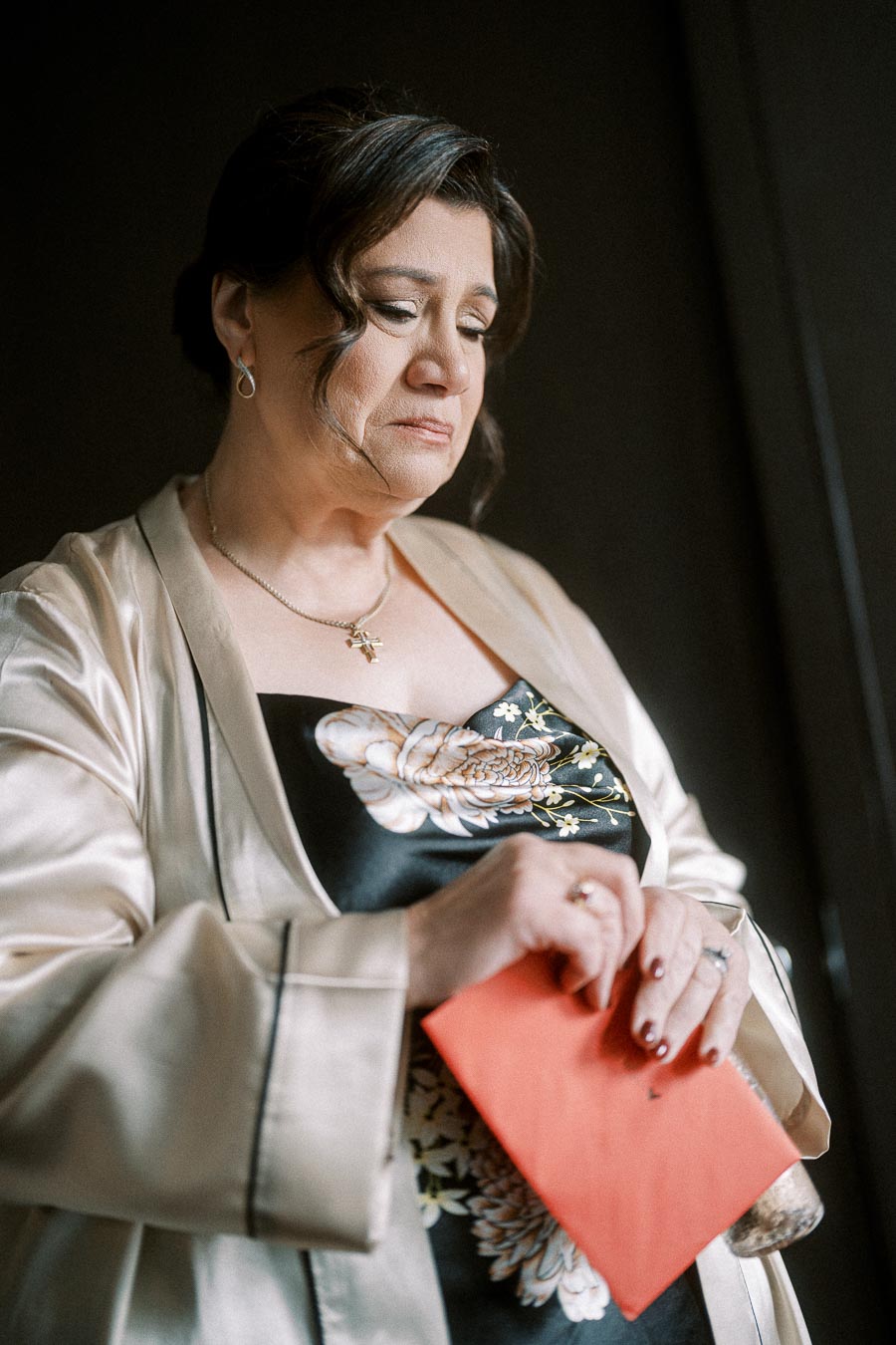 A woman in an elegant beige satin robe with floral patterns holds a red envelope, looking contemplative.