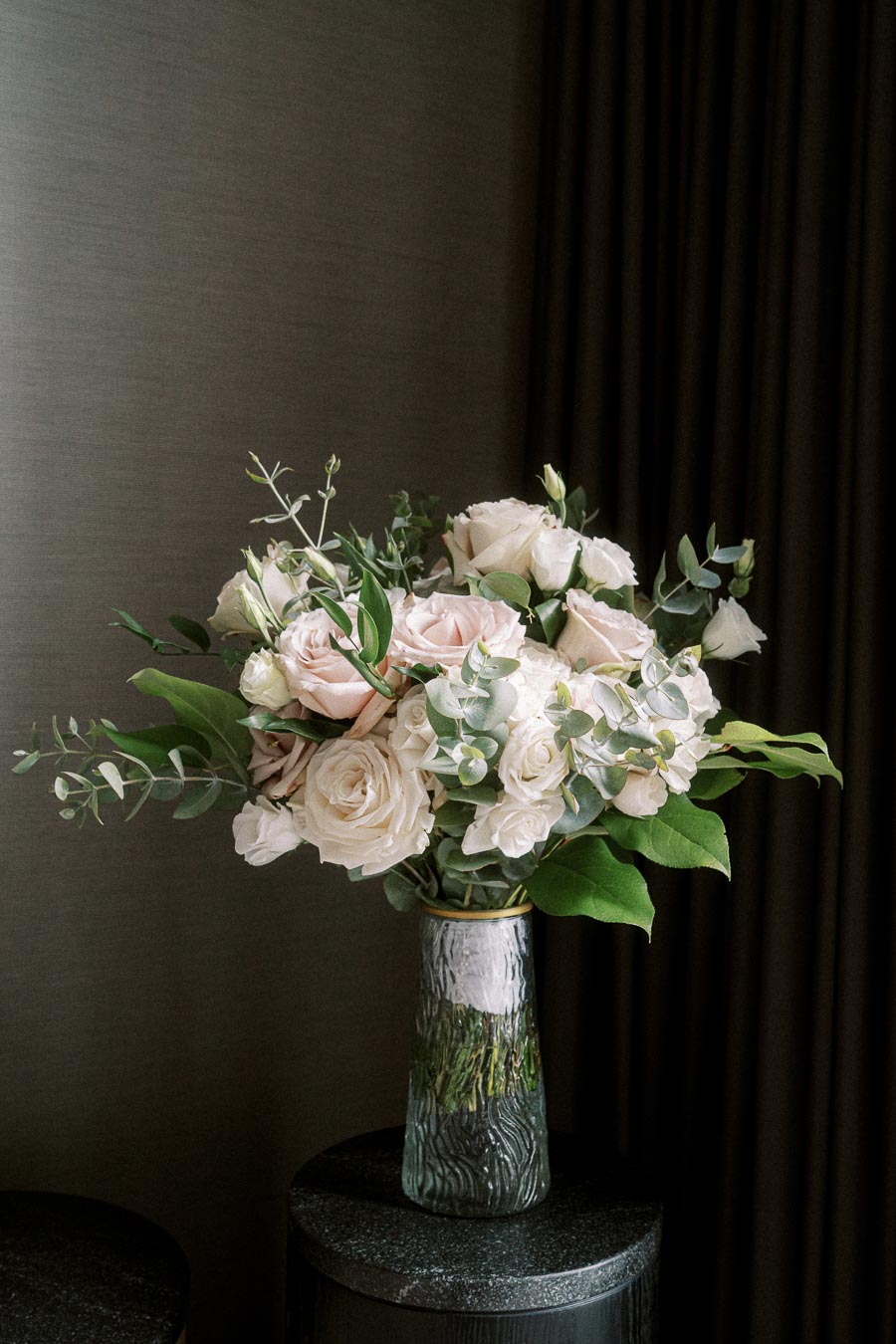 A bouquet of elegant white and blush pink roses with green foliage in a textured glass vase, placed on a dark round table