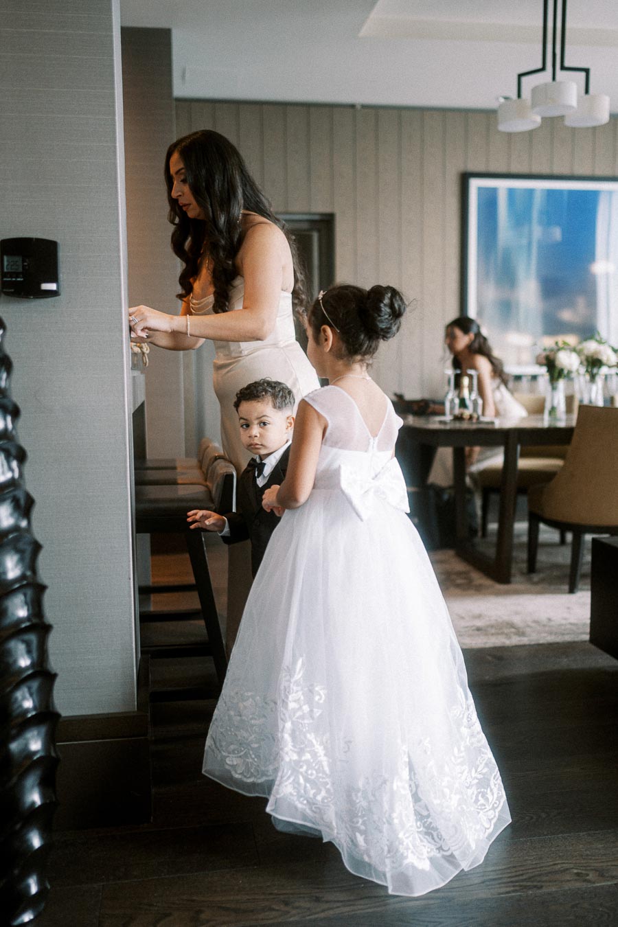 A woman in an elegant dress stands at a counter, while two children dressed formally—a girl in a white dress with a bow and