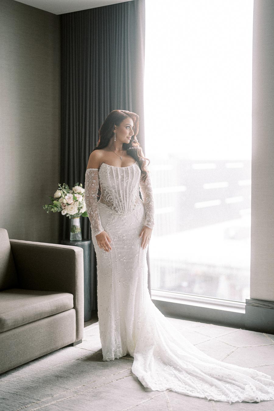 Bride in elegant white wedding dress with lace details stands gracefully by a large window, soft light illuminating her long