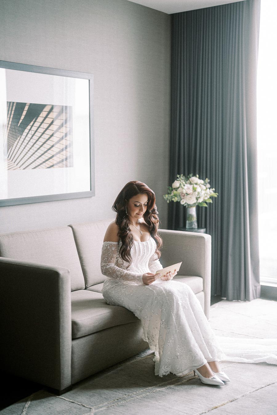 Bride wearing an elegant white off-shoulder wedding dress, sitting on a couch in a softly lit room, reading a card; elegant