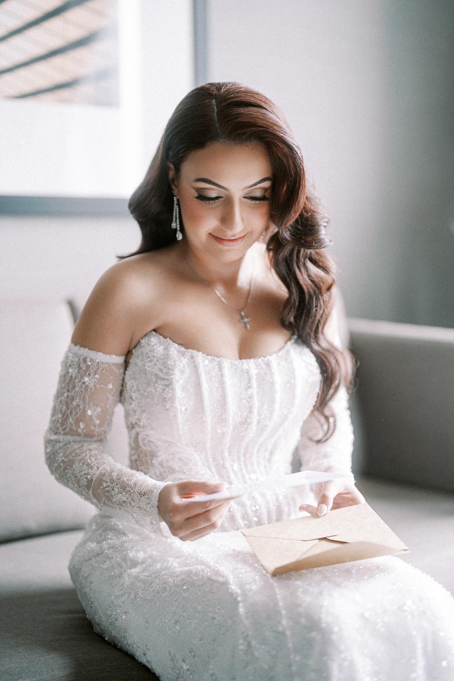 Bride in an off-the-shoulder lace wedding dress reading a letter while sitting on a sofa, showcasing elegant bridal fashion