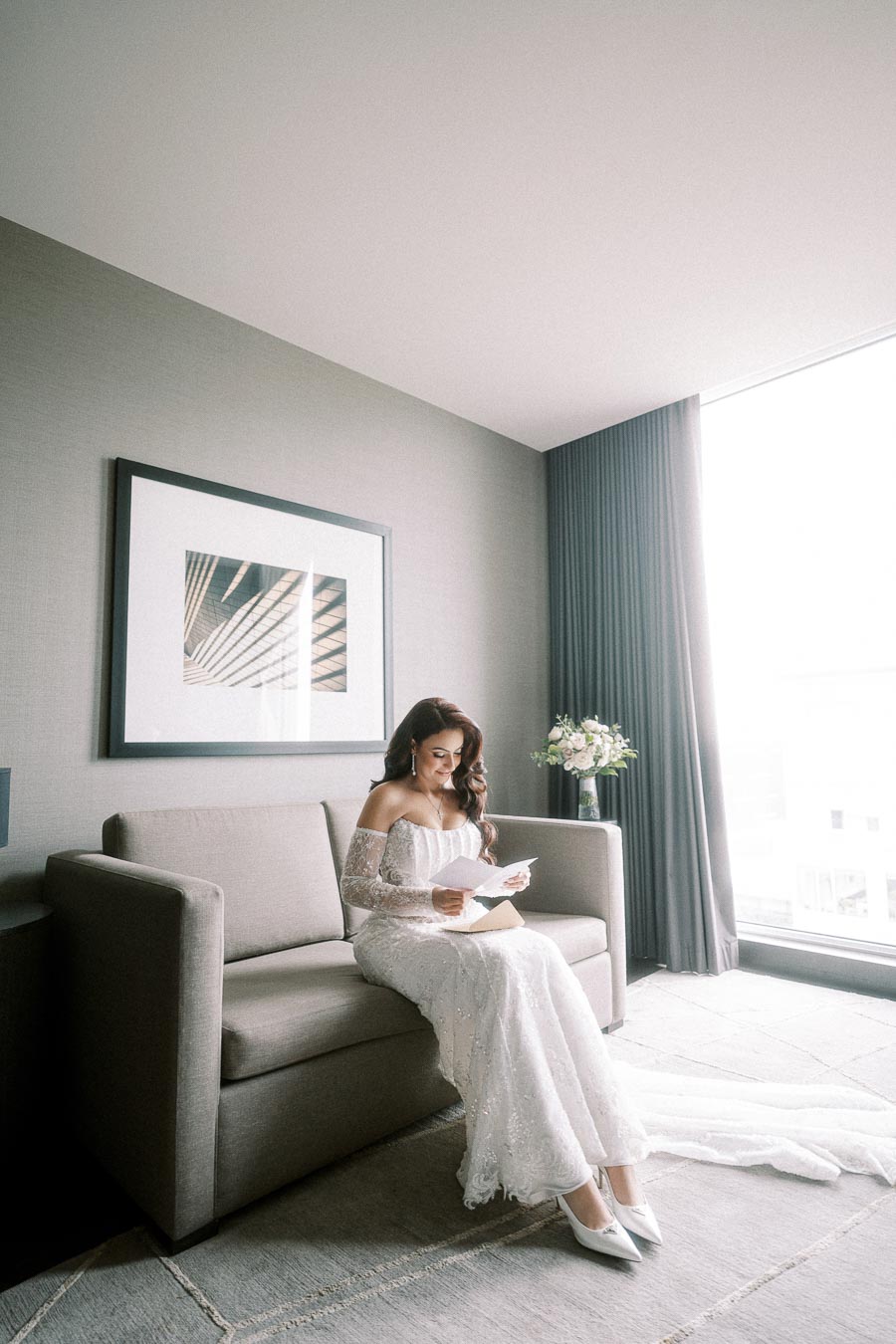 Bride in elegant white wedding gown reading a letter while seated on a modern sofa in a bright room with large window and