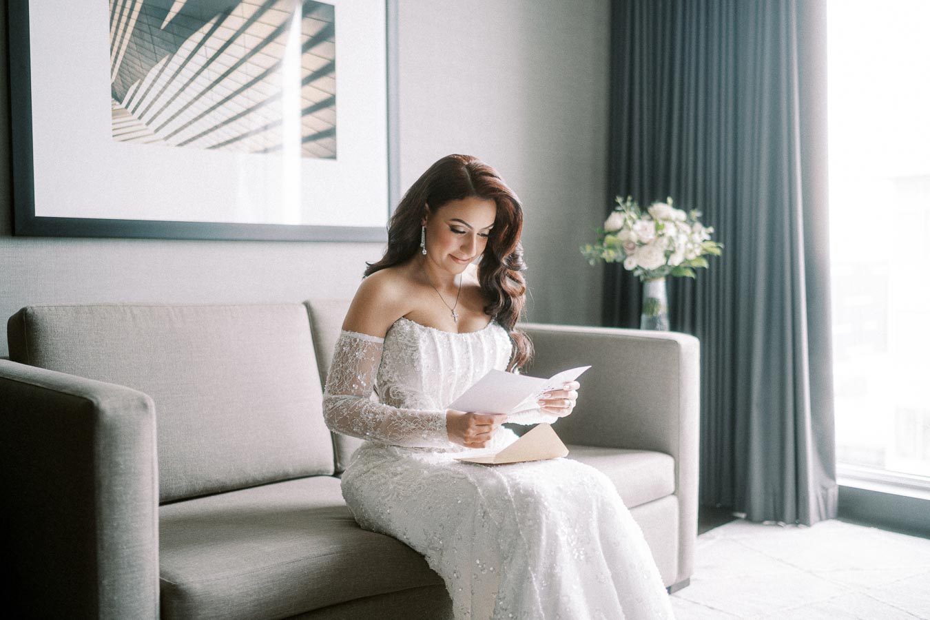 Bride in elegant lace wedding dress reading a letter on a sofa, with natural light coming through the window and a bouquet
