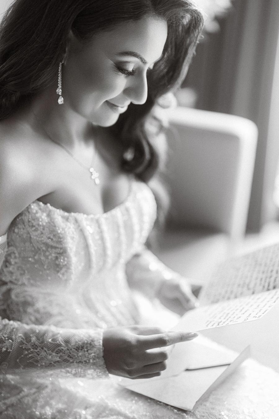 A bride in an elegant lace wedding dress smiles while reading a heartfelt letter before her wedding ceremony.