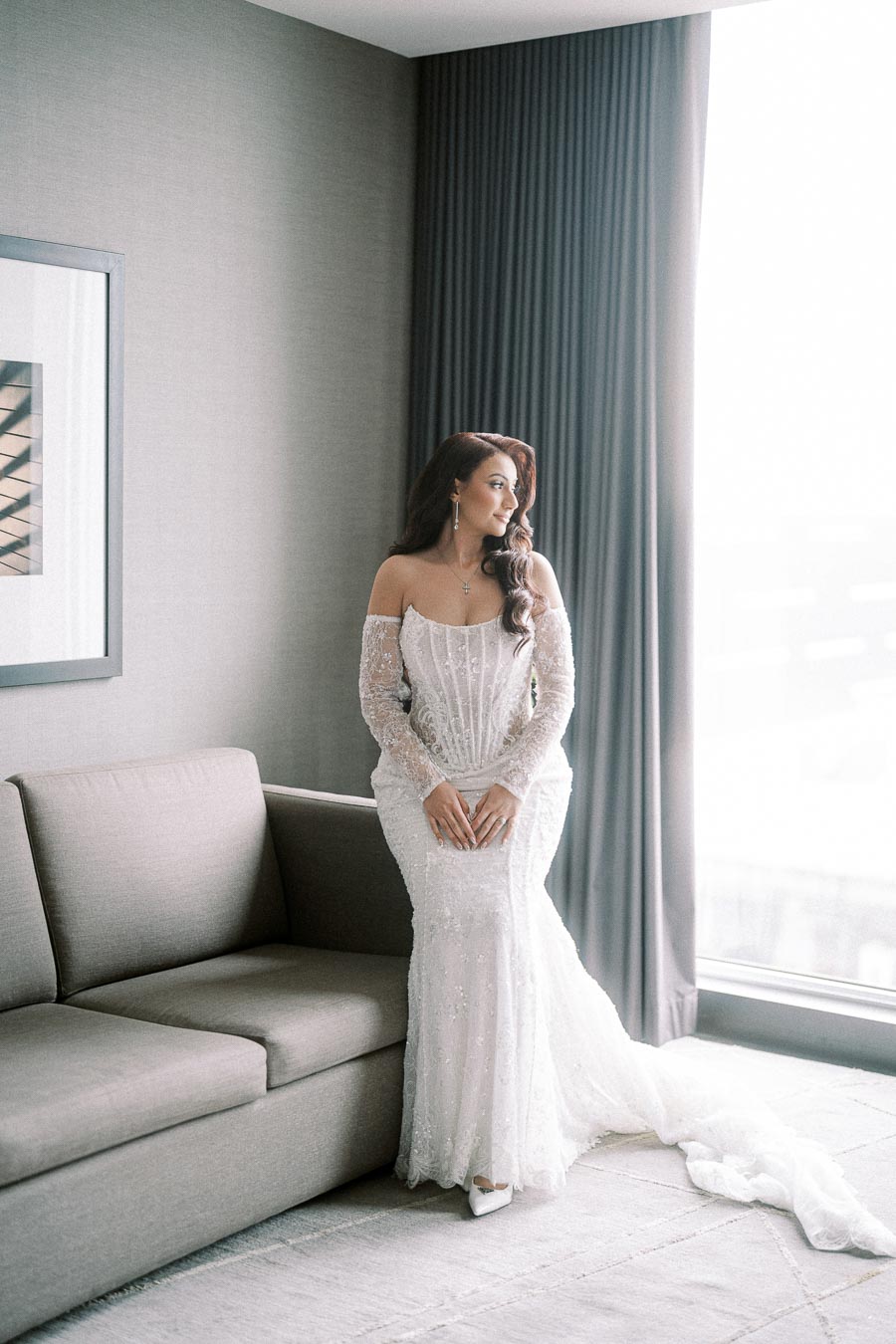 Bride in an elegant, lace off-shoulder wedding gown poses by a window in a modern room, exuding timeless beauty and