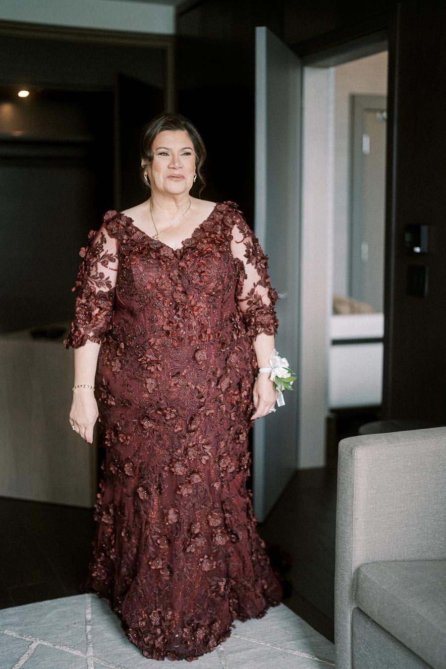 Elegant woman wearing a burgundy floral lace gown, standing in a stylish room with modern decor.