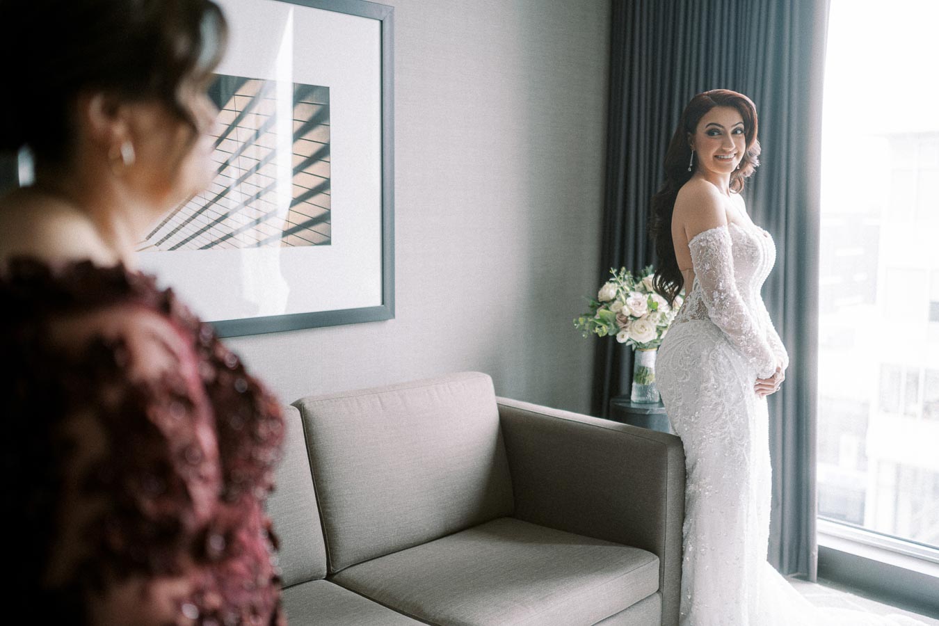 A bride in an elegant lace wedding gown stands by a window, with soft natural light illuminating her smile. A bouquet of