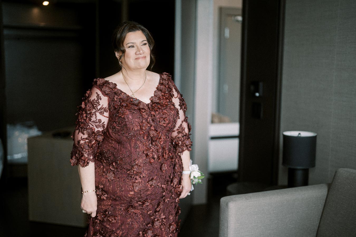 A woman wearing a detailed burgundy lace dress stands in a modern room, smiling as she looks ahead. The elegant dress