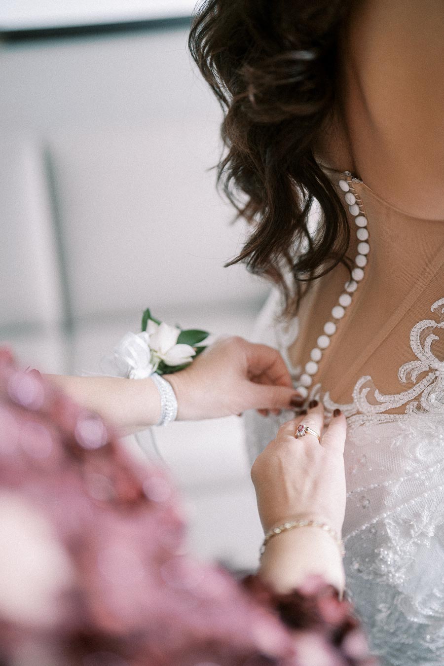 A close-up of a bride in a lace wedding dress being buttoned up by a person wearing a purple outfit and wrist corsage,