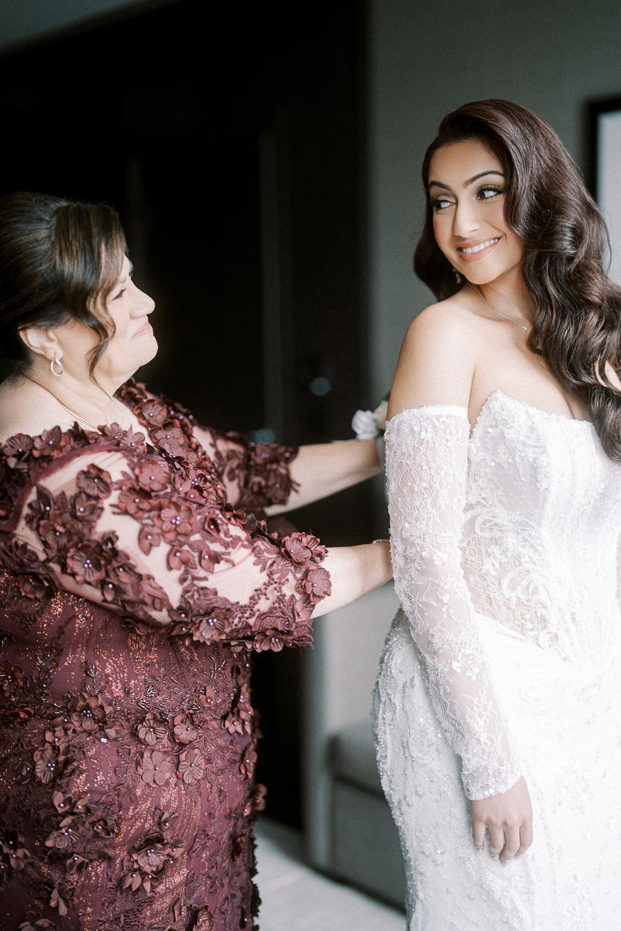 Bride in a white lace wedding gown smiling, while an older woman in an embroidered burgundy dress adjusts her gown,