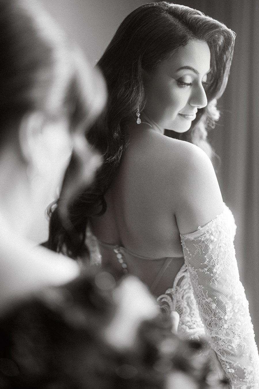 Black and white photograph of a bride in an elegant lace wedding dress, looking over her shoulder with soft curls framing