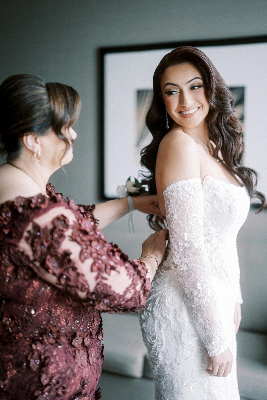 A bride in a lace wedding dress smiles as a woman in a burgundy gown helps with the dress fitting before the ceremony.