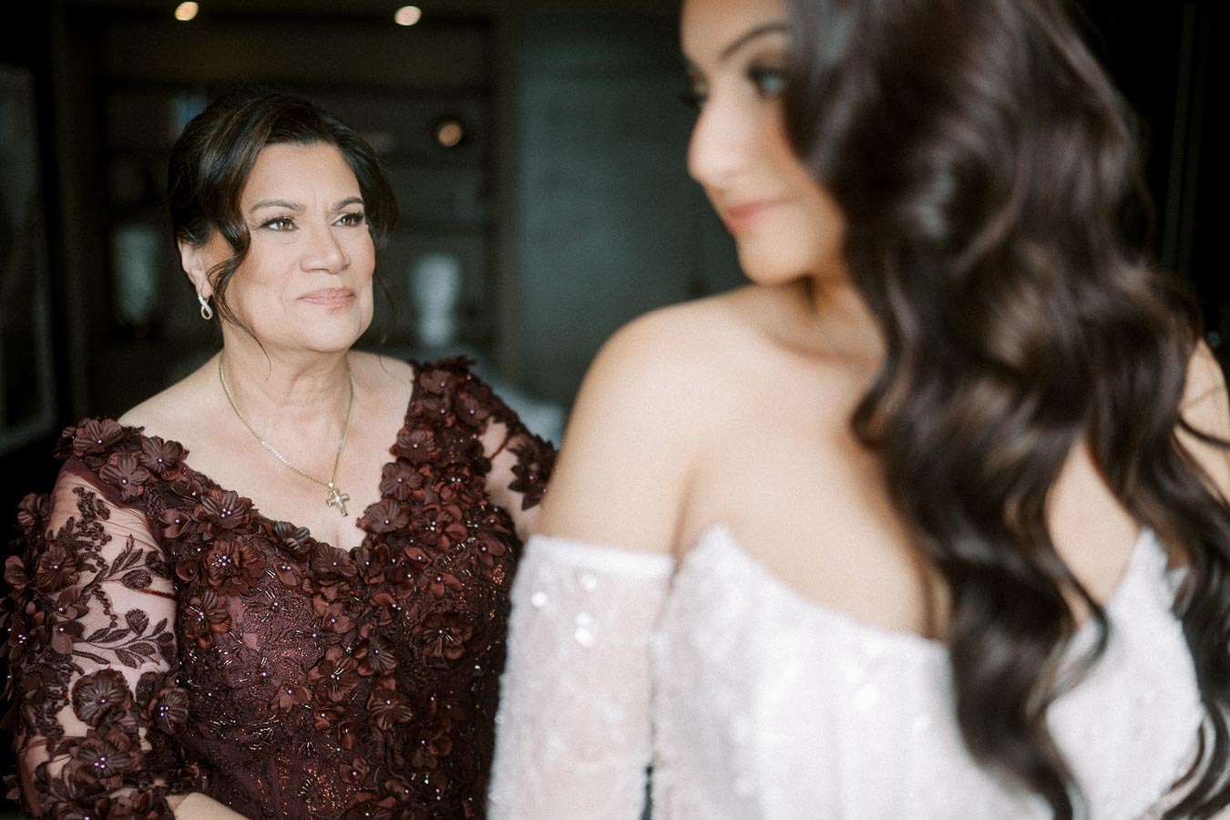 A woman in an elegant floral dress smiles warmly at a bride with long, flowing hair in an off-the-shoulder wedding gown,