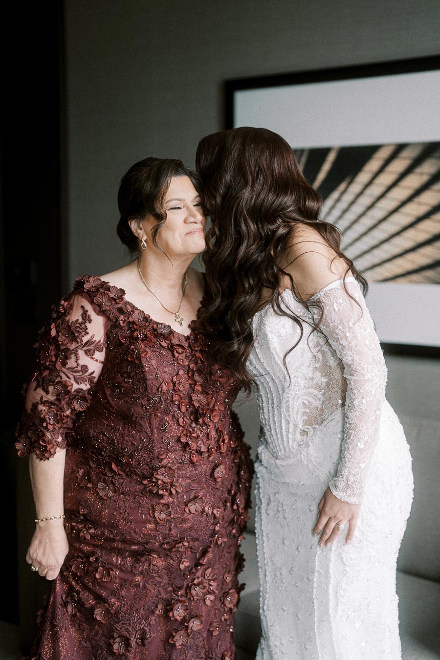 A bride in a white lace wedding gown shares a loving moment with her mother, who is wearing an elegant dark red floral dress.